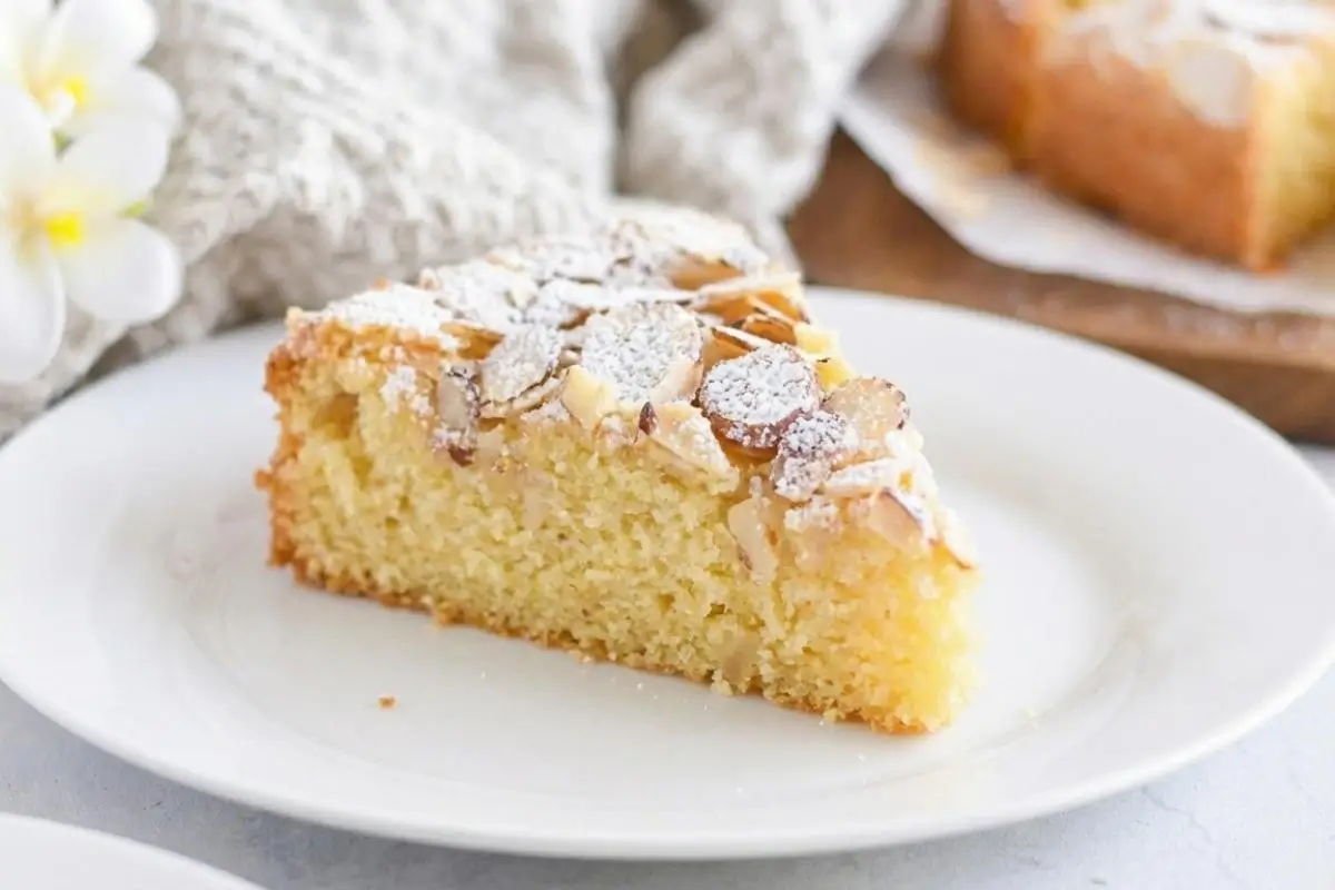 How To Store Your Italian Almond Cake A close-up view of a moist slice of Italian almond cake on a white plate, generously topped with toasted sliced almonds and dusted with powdered sugar, with a soft linen cloth in the background.