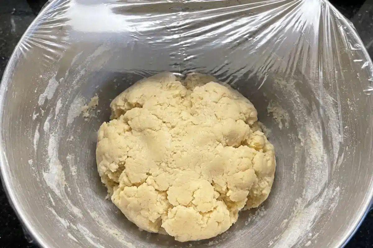 How to Make Apricot Cream Cheese Cookies Step by Step A ball of freshly mixed dough for apricot cream cheese cookies resting in a silver mixing bowl, covered tightly with clear plastic wrap to chill.