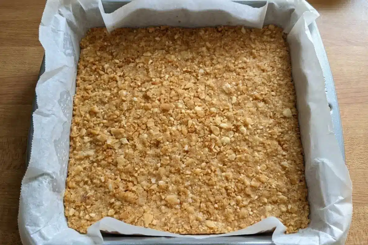 How to Make Cannoli Cheesecake Bars Step by Step A top-down view of a golden, buttery crumb crust pressed firmly and evenly into a parchment-lined square baking pan, illustrating the simple first step in how to make cannoli cheesecake bars.