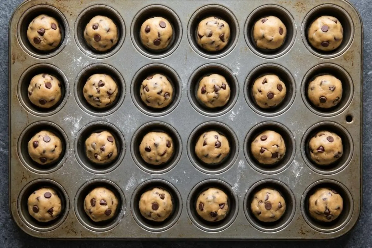 How to Make Cannoli Cookie Cups An overhead view showing a key step in how to make cannoli cookie cups, featuring a wonderfully weathered metal mini muffin pan filled with perfectly portioned, round balls of chocolate chip cookie dough.