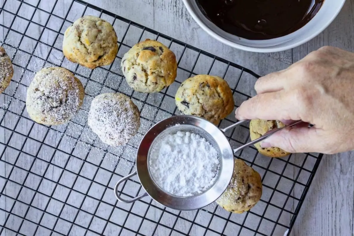 How to Make Cannoli Cookies An overhead view showing a beautiful finishing step in how to make cannoli cookies, featuring a hand using a small metal sieve to gently dust white powdered sugar over soft, freshly baked cookies on a black wire cooling rack.