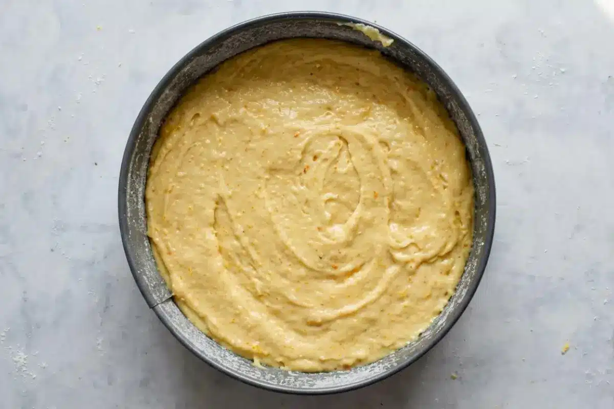 How to Make Italian Almond Cake A top-down view of smooth, pale yellow Italian almond cake batter spread evenly inside a round metal springform pan on a rustic, lightly flour-dusted surface.