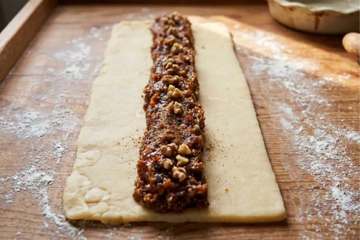How to Make Italian Fig Cookies A rustic wooden cutting board dusted with flour showing a step in how to make Italian fig cookies, featuring a rectangle of pale rolled dough with a thick line of dark, rich fig filling, topped with chopped walnuts and spices, laid down the center.