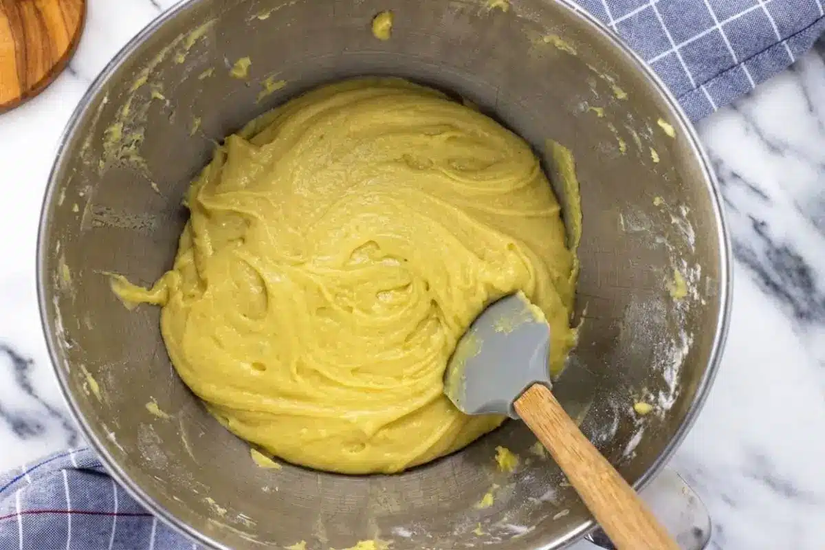 How to Make Italian Pizzelle Cookies An overhead view showing a key step in how to make Italian pizzelle cookies, featuring a smooth, thick, golden batter freshly mixed in a stainless steel bowl with a grey silicone spatula resting inside.