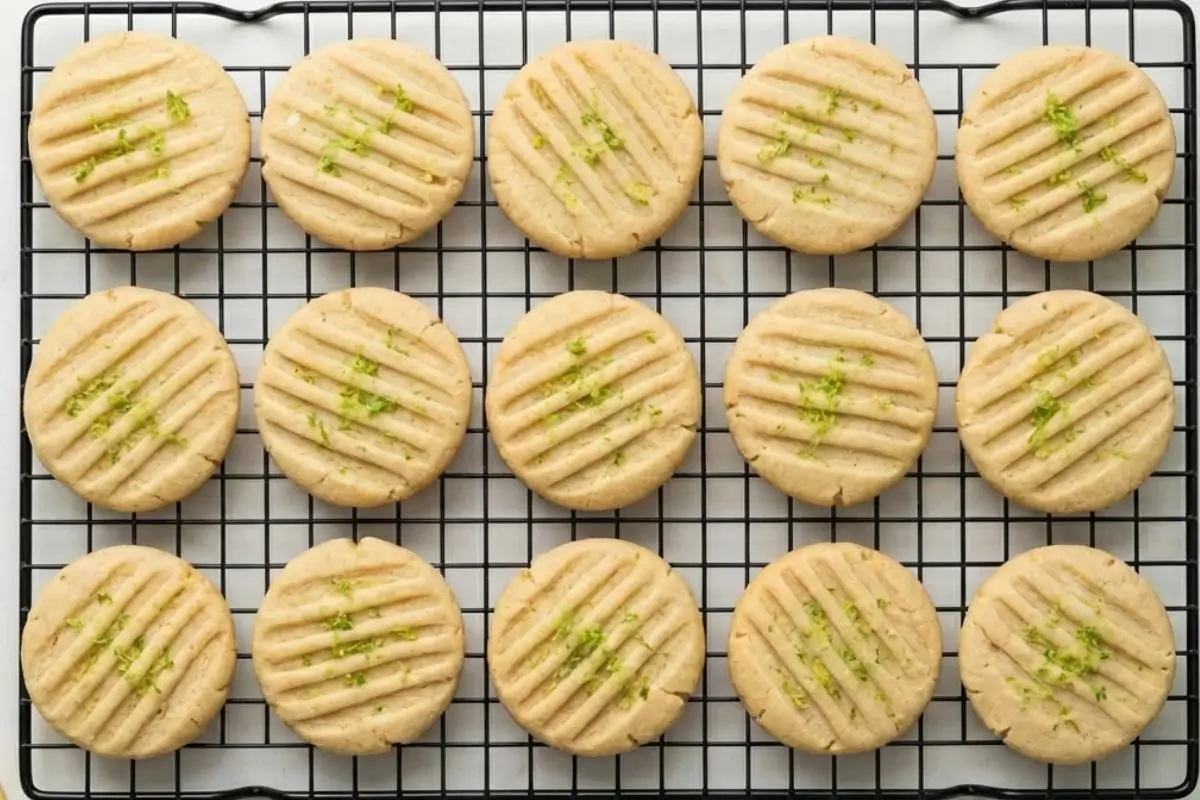 How to Make Lemon Lime Basil Italian Shortbread Cookies Step-by-Step Neat rows of freshly baked lemon lime basil Italian shortbread cookies, featuring rustic pressed fork marks and sprinkled with fresh lime zest, resting on a black wire cooling rack.