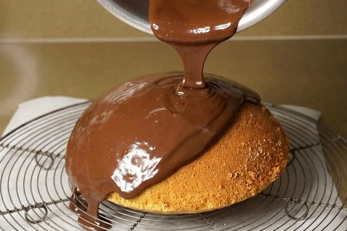 How to Make Parrozzo A thick, glossy chocolate glaze being poured from a saucepan over a freshly baked golden dome cake on a wire cooling rack, showing the final beautiful step of how to make parrozzo.