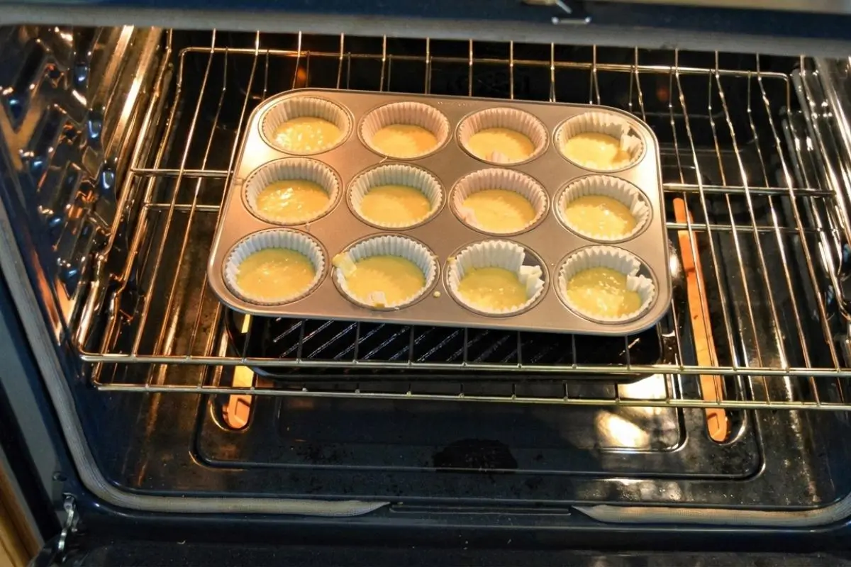 How to Make Tiramisu Cupcakes A twelve-cup metal muffin tin filled with light yellow cake batter in white paper liners sits on a wire rack inside a warm oven, illustrating the baking step of how to make tiramisu cupcakes.