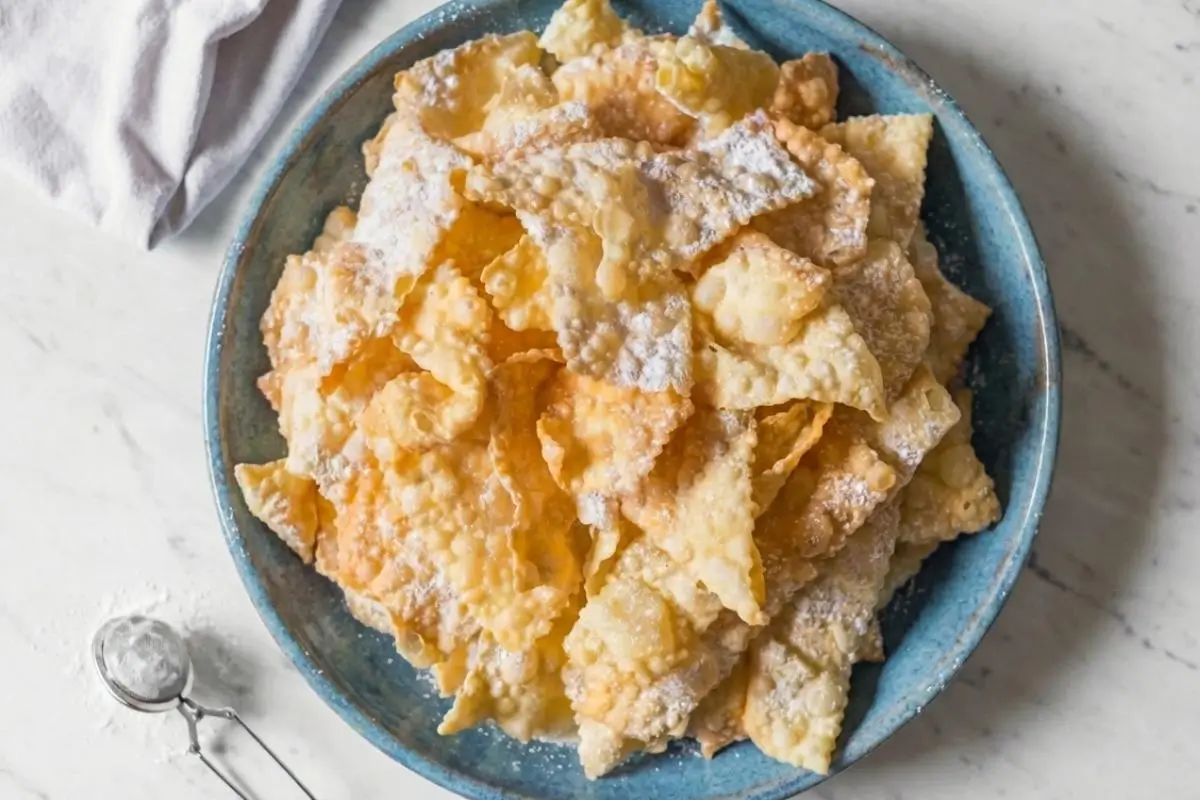 Modern Plating & Holiday Entertaining fot Italian Crostoli An overhead view showing a beautiful way of serving Italian crostoli, featuring a large, rustic blue ceramic bowl piled high with golden, crispy fried pastries generously dusted with powdered sugar, sitting on a white marble counter next to a small metal sugar dusting wand.