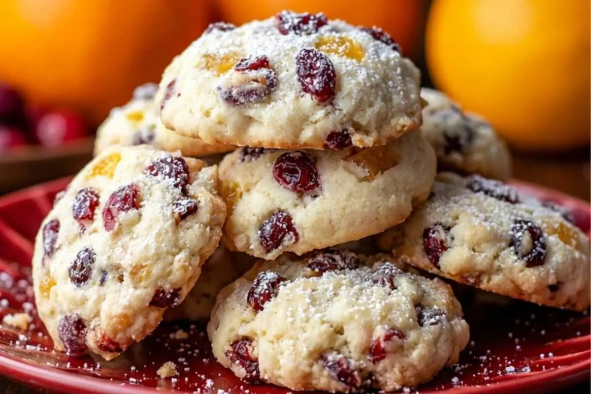 Orange Cranberry Ricotta A close-up view of a stack of freshly baked orange cranberry ricotta cookies lightly dusted with powdered sugar, resting on a decorative red plate with fresh oranges and cranberries softly blurred in the background.