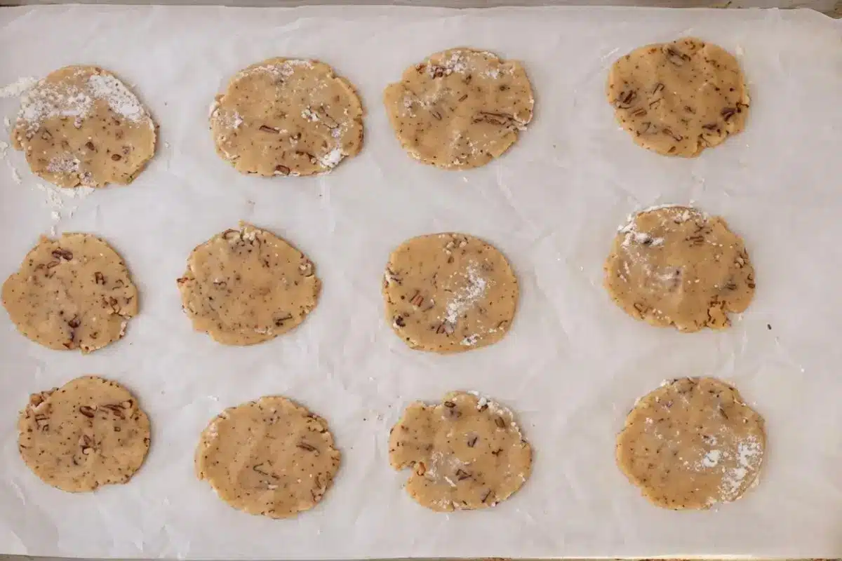Rena's Cookie Rescue Guide for Chewy Brown Sugar Pecan Oatmeal Cookies An overhead view of twelve round disks of raw chewy brown sugar pecan oatmeal cookie dough resting on white parchment paper, speckled with nuts and showing a light, rustic dusting of flour on top.