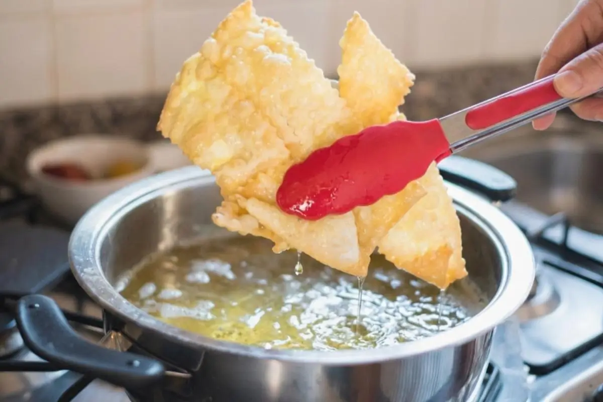 Rena's Frying Masterclass fot Italian Crostoli A close-up, action shot showing the process of frying Italian crostoli, featuring bright red silicone tongs lifting a cluster of large, golden, beautifully bubbled pastries out of a stainless steel pot of hot oil on a stovetop.