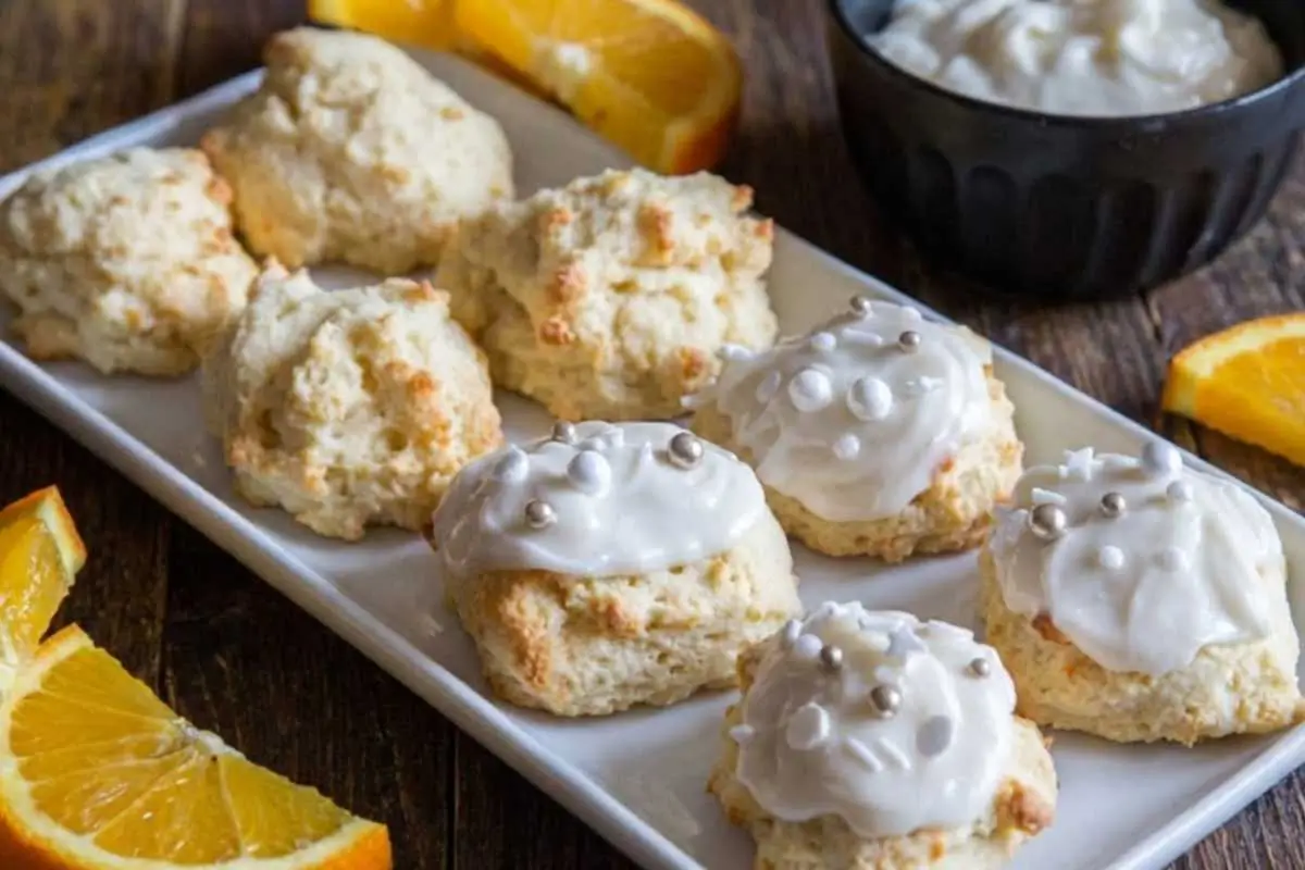 Rena's No-Fail Baking Secrets for Italian Orange Ricotta Cookies A white rectangular platter showcasing both fluffy, unglazed bakes and fully decorated no fail Italian orange ricotta cookies topped with sweet white icing and silver sprinkles, resting on a rustic wooden table with fresh orange slices.