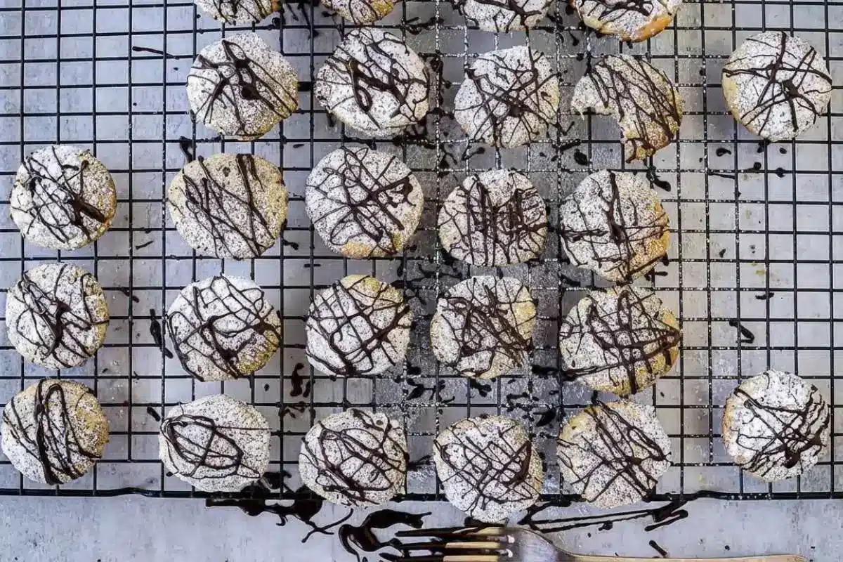 Rena's No Fail Tips For Cannoli Cookie An overhead view of freshly baked, no fail cannoli cookies resting on a black wire cooling rack, generously dusted with powdered sugar and decorated with a rustic dark chocolate drizzle, with a chocolate-covered fork resting at the bottom and one cookie showing a bite taken out of it.
