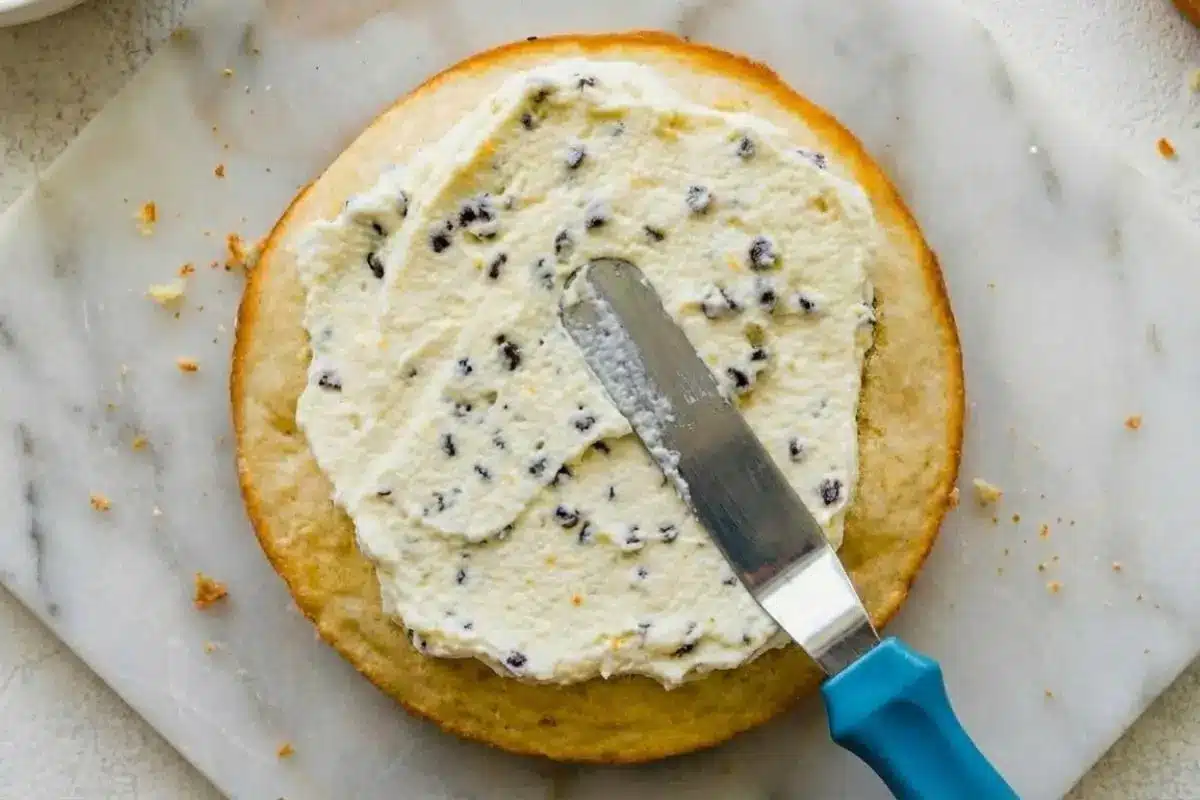 Rena’s No Fail Tips For Cannoli Cream Cake Step by Step An overhead view showing one of the best cannoli cream cake tips: using an offset spatula with a bright blue handle to gently spread a thick, chocolate chip-studded ricotta filling evenly over a golden sponge cake layer resting on a rustic marble slab.