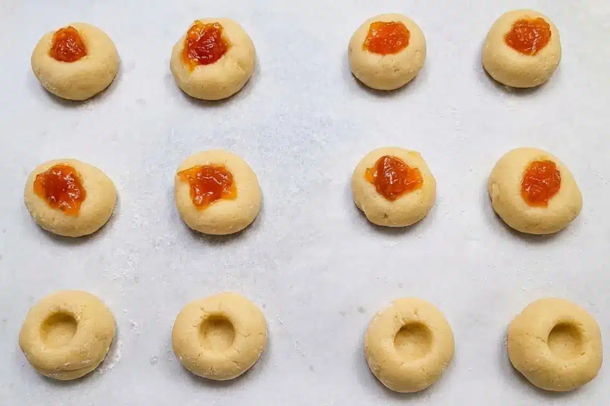 Rena's No-Fail Tips For Your Apricot Cream Cheese Cookies A top-down view of unbaked apricot cream cheese cookies on a lined baking sheet, showing the step-by-step process of making small thumbprint indentations in the dough balls and filling them with bright orange apricot preserves.