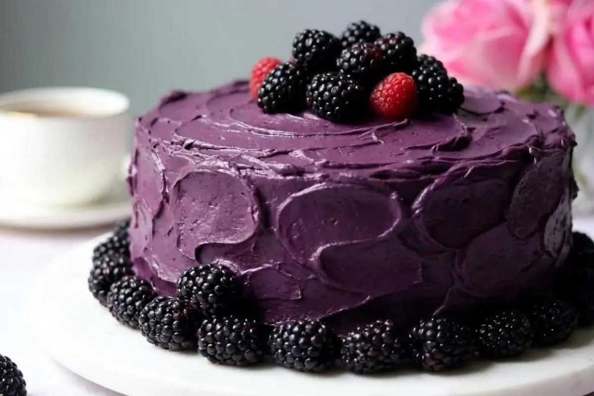 Rena's No-Fail Tips For Your Dreamiest Blackberry Velvet Cake A gorgeous whole blackberry velvet cake covered in rustic, swooping dark purple frosting and adorned with fresh blackberries and raspberries, sitting on a white serving platter with a teacup and pink roses in the background.