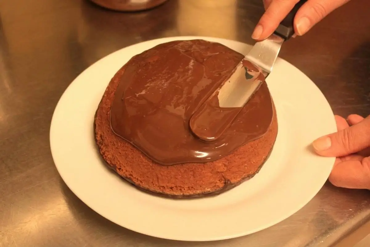 Rena's No-Fail Tips For Your Parrozzo A pair of hands using a metal offset spatula to gently smooth a rich, glossy chocolate glaze over a rustic baked almond cake on a simple white plate, demonstrating no-fail tips for your parrozzo.