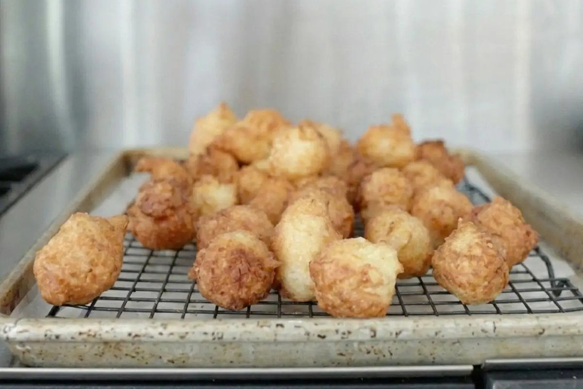 Rena's No-Fail Tips For Your Zeppole A well-loved, rustic baking sheet fitted with a black wire cooling rack, holding a fresh batch of golden brown, irregularly shaped fried dough, demonstrating no-fail zeppole tips in a home kitchen.