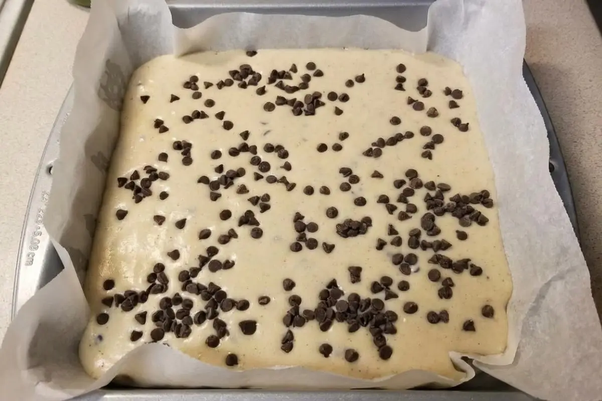 Rena's No Fail Tips The Science of Success A top-down view of smooth, unbaked ricotta batter heavily sprinkled with mini chocolate chips inside a parchment-lined square baking pan, demonstrating our no fail cannoli cheesecake tips for a perfectly even bake.