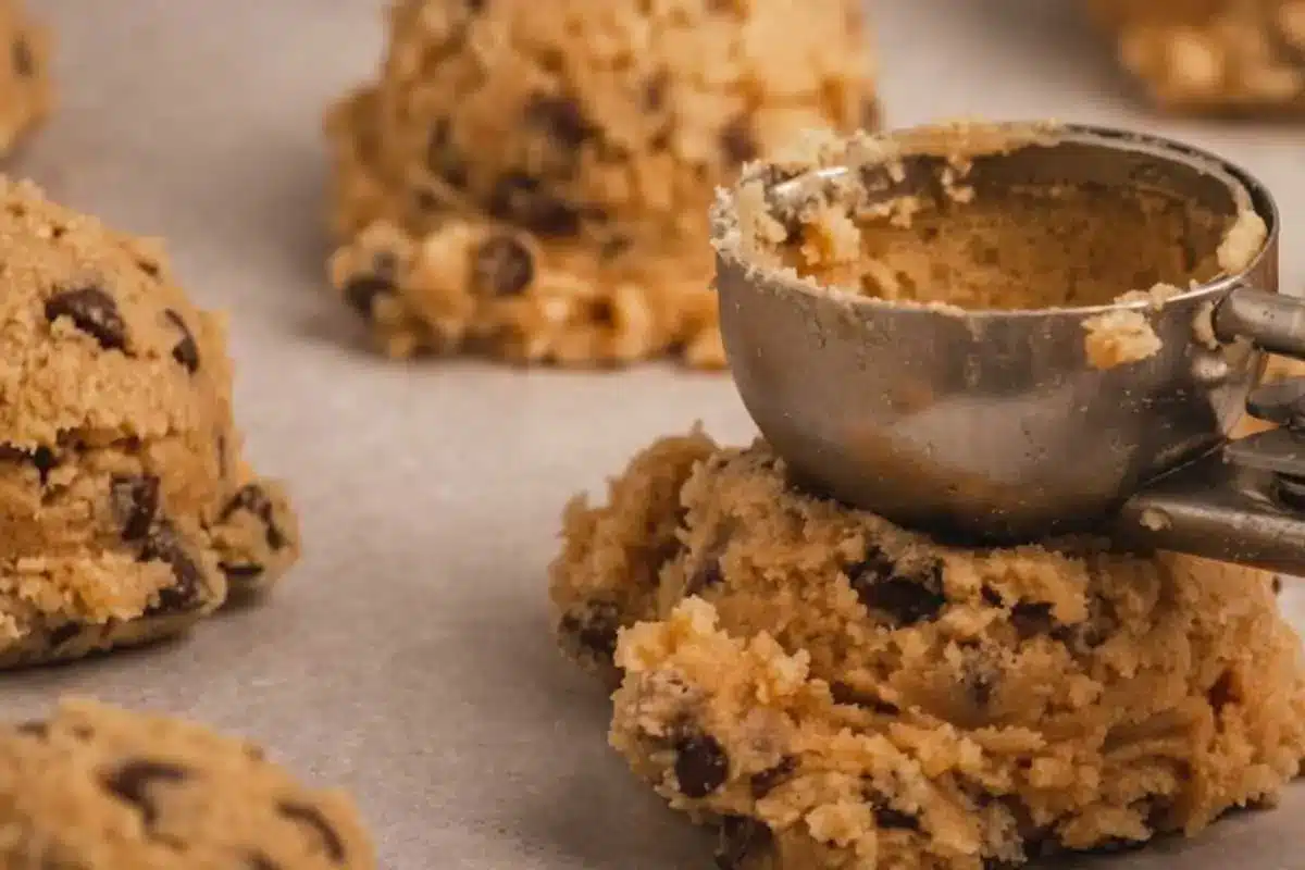 Rena's Ricotta Rescue A close-up view of soft ricotta cannoli cookie dough studded with mini chocolate chips, being released from a metal cookie scoop in rustic mounds onto a parchment-lined baking sheet.