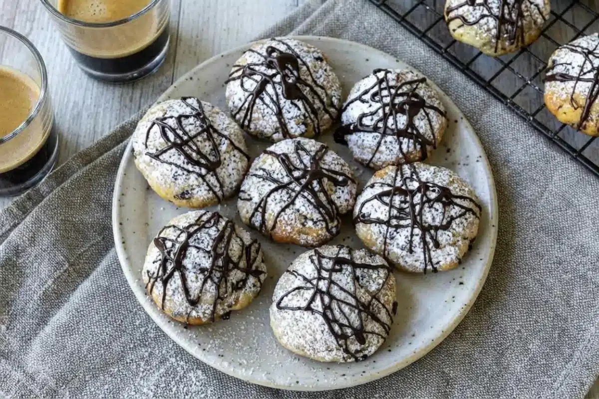 Storage & Freezing For Cannoli Cookies Recipe An overhead view showing how to store cannoli cookies, featuring a rustic ceramic plate filled with powdered and chocolate-drizzled cookies resting on a warm grey linen napkin next to two small glasses of fresh espresso.