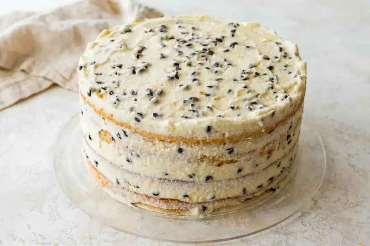 Storage & Freezing The Pro Tip for Busy Hosts A fully assembled, four-layer naked dessert resting on a clear glass cake plate next to a rustic linen napkin, demonstrating the ideal, unfrosted stage for storing cannoli cream cake in the fridge or freezer.