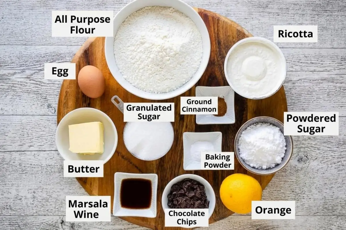 The Foolproof Ingredients List For Your Cannoli Cookie Recipe An overhead view of foolproof cannoli cookie ingredients arranged in small white bowls on a rustic round wooden board, featuring flour, fresh ricotta, butter, chocolate chips, a whole orange, and a splash of Marsala wine.