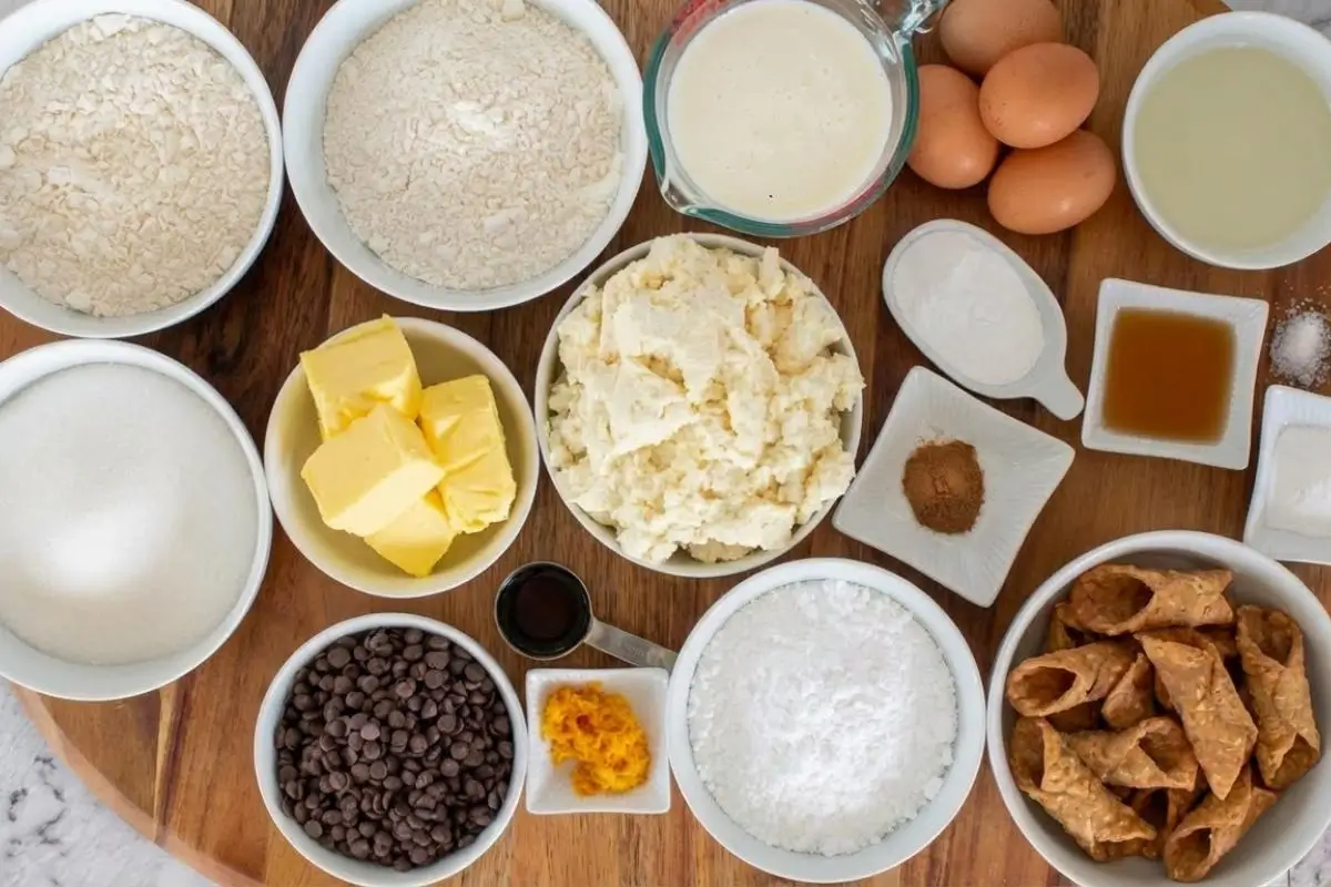 The “Foolproof” Ingredients List For Your Cannoli Cream Cake An overhead shot of foolproof cannoli cream cake ingredients gathered on a rustic wooden board, featuring white bowls filled with fresh ricotta, butter, mini chocolate chips, flour, sugar, eggs, and crispy broken cannoli shells.