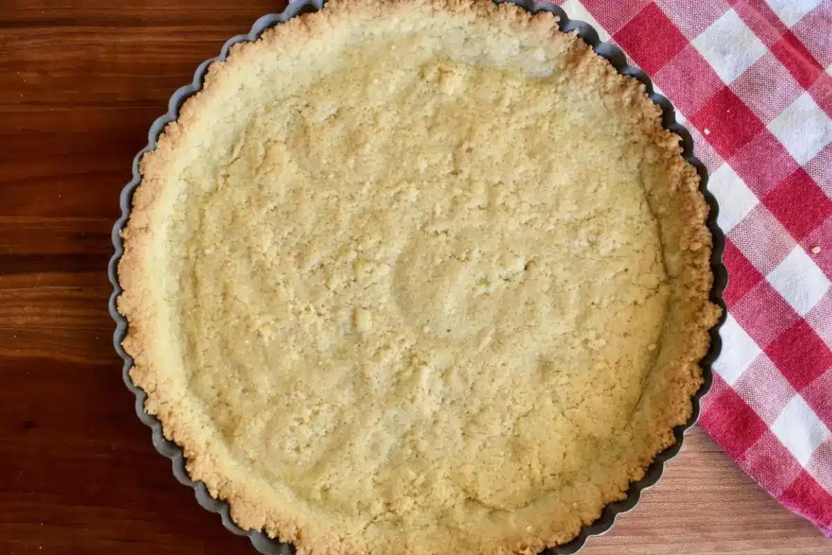 The Foolproof Ingredients List For Your Ricotta Crostata An overhead view of a simple, buttery shortcrust pastry pressed into a metal fluted tart pan, sitting on a warm wooden table next to a red and white checkered napkin, ready for its ricotta filling.