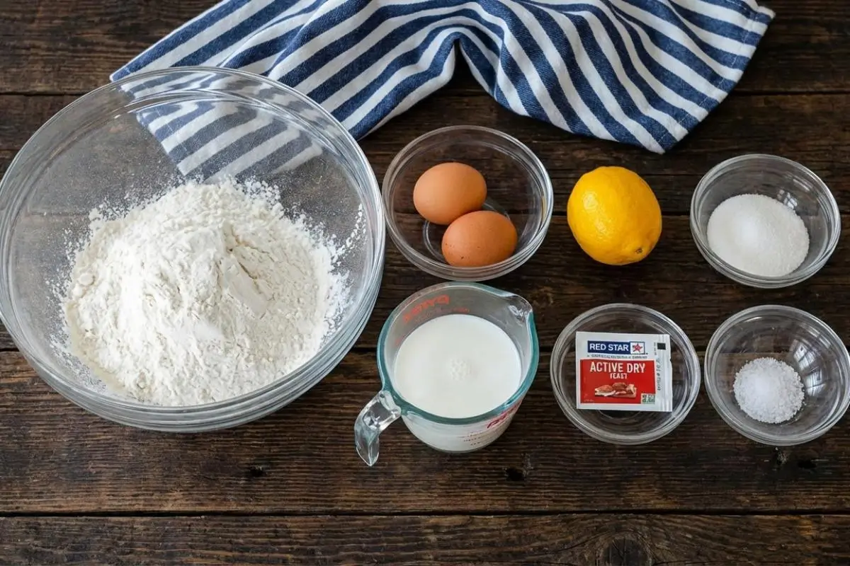 The Foolproof Ingredients List For Your Zeppole A rustic wooden table displaying foolproof zeppole ingredients in simple glass bowls, including flour, fresh eggs, a whole lemon, milk, sugar, salt, and active dry yeast, arranged next to a blue and white striped kitchen towel.