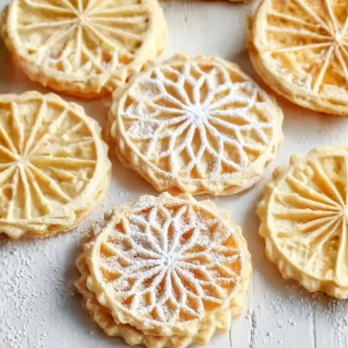 Perfect Italian Pizzelle Cookies for Sweet Memories Italian Pizzelle cookies An overhead view of delicate, round traditional Italian holiday cookies with intricate star and wagon-wheel patterns, some lightly dusted with powdered sugar, scattered across a rustic white wooden table.