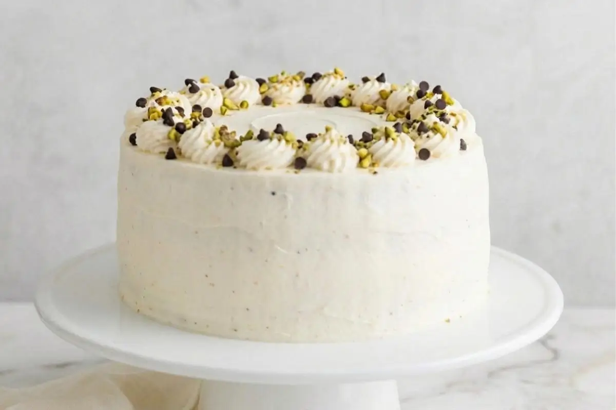 Why This Cannoli Cake with Chocolate Chips Works A beautifully frosted white cannoli cake with chocolate chips, elegantly decorated with piped ricotta rosettes, crushed pistachios, and mini chocolate chips on a classic white cake stand.