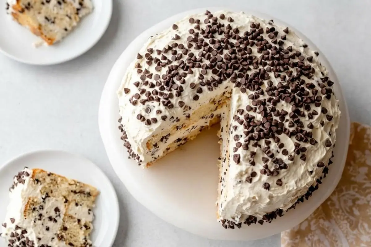 Why This Cannoli Cream Cake Works An overhead view of a sliced homemade cannoli cream cake on a white cake stand, showing three fluffy cake layers sandwiched with sweet ricotta filling and covered in mini chocolate chips, with two generous slices ready to serve on plates nearby.