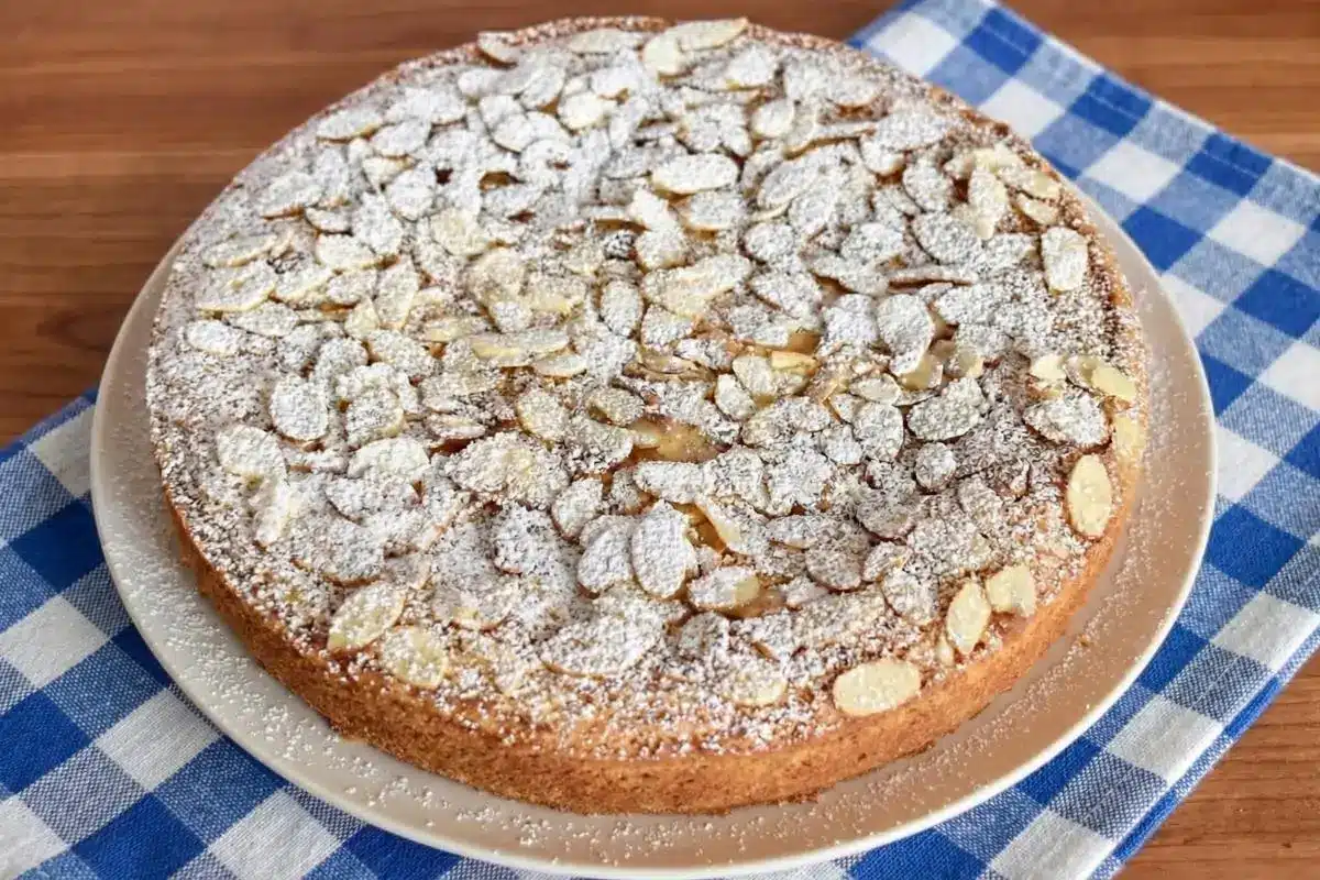 Why You Will Fall In Love With This Italian Almond Cake A freshly baked Italian almond cake heavily dusted with sweet powdered sugar and topped with flaked almonds, sitting on parchment paper and a wooden board with a slice beautifully cut out.