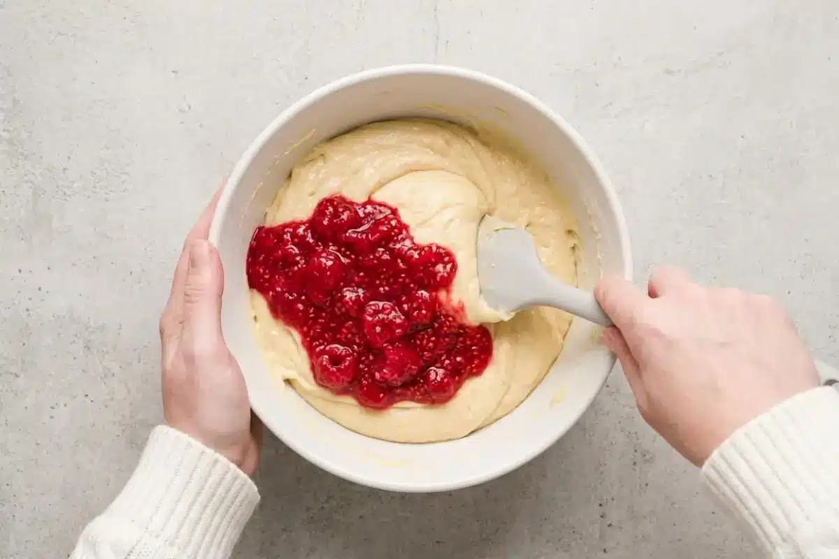 Fold the Fruit For Raspberry Cake An overhead view of a baker wearing a cozy cream sweater using a grey silicone spatula for gently folding raspberries into cake batter inside a large white ceramic bowl.