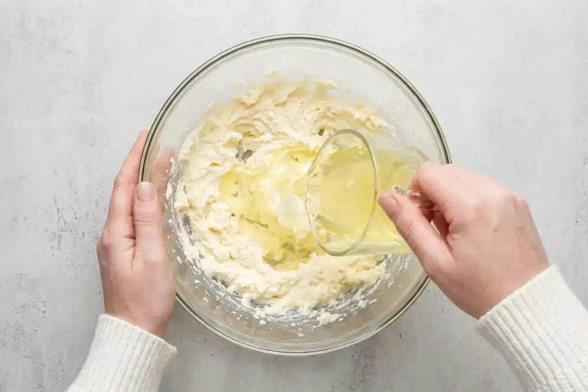 How to Make Classic White Almond Cake Step-by-Step An overhead view of a baker's hands wearing a cozy white sweater, gently pouring wet ingredients from a glass measuring cup into a large glass bowl of creamed butter to make a classic white almond cake batter.