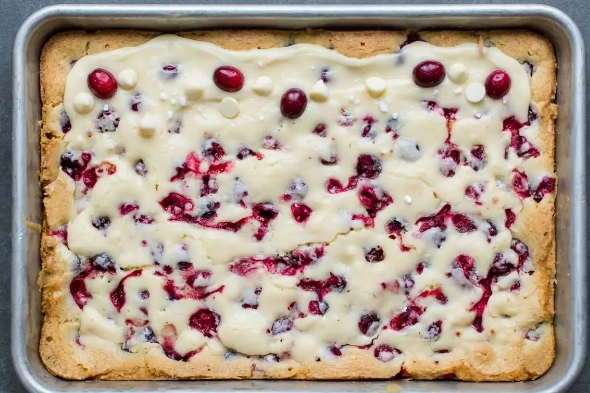 How to Make Cranberry Cookie Bars Step-by-Step A top-down view of cranberry cookie bars in a silver baking pan, showing the golden cookie base topped with a creamy white chocolate swirl and fresh whole cranberries.