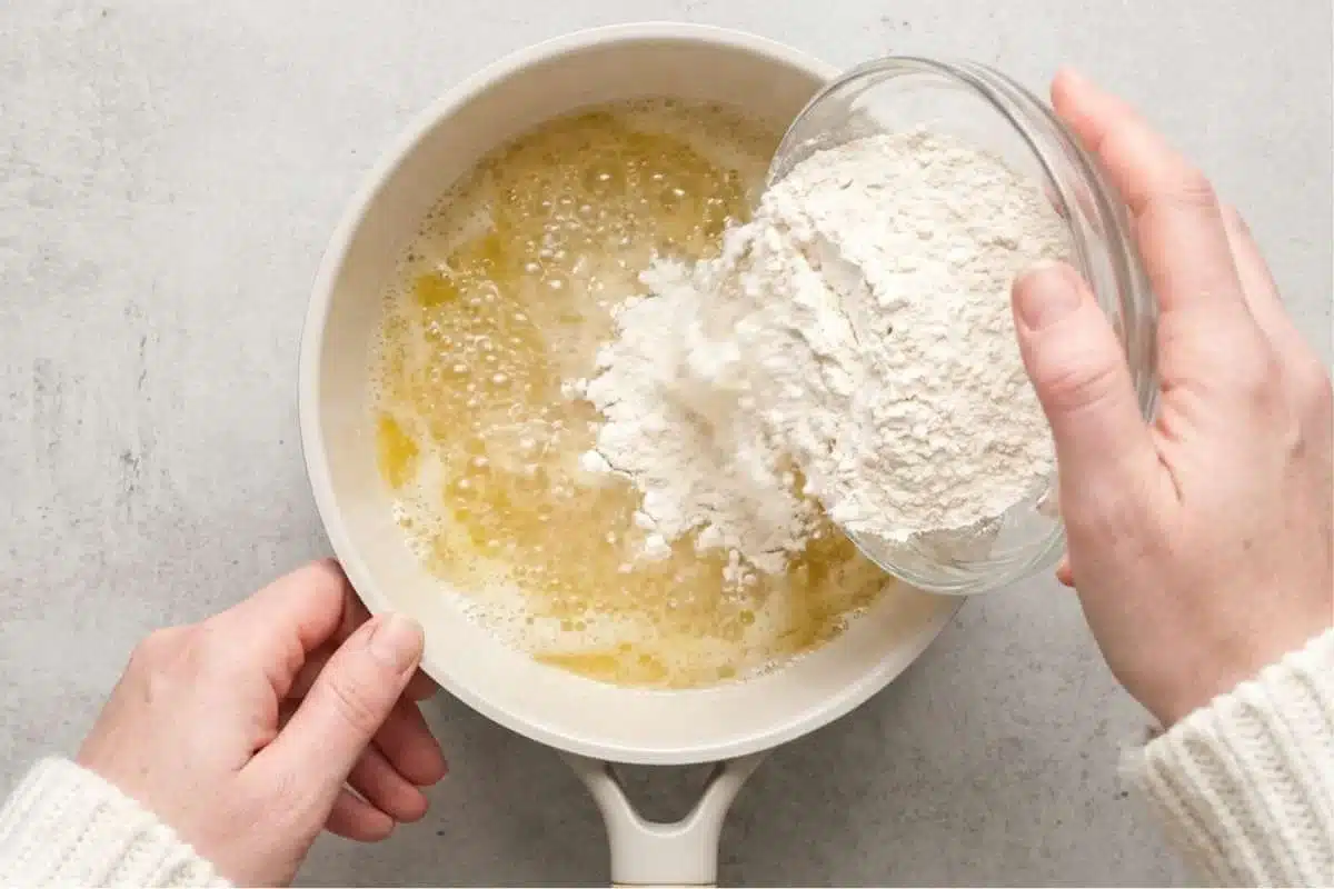 How to Make Cream Puffs Step-by-Step (1) Flour being added to a butter and egg mixture in a saucepan while making Mom’s famous cream puffs recipe from scratch.
