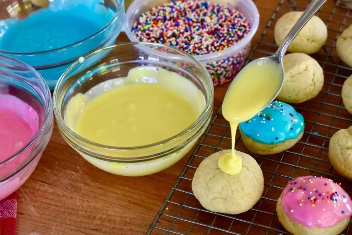 How to Make Easter Anisette Cookies A close-up showing how to make Easter anisette cookies, featuring a silver spoon gently drizzling smooth, pastel yellow glaze over a soft, round cookie on a wire cooling rack, with bowls of bright blue and pink icing and rainbow sprinkles nearby on a wooden table.