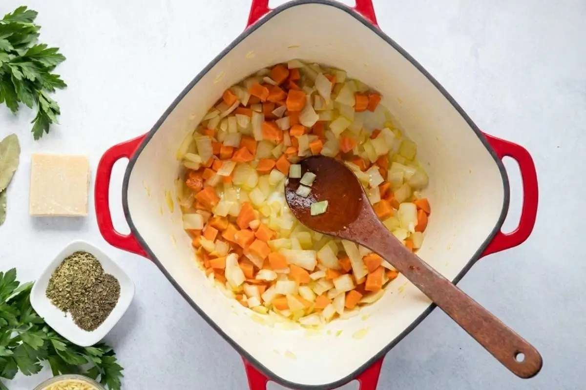 How to Make Italian Meatball and Orzo Soup Step-by-Step An overhead view showing how to make Italian meatball and orzo soup, featuring chopped onions and carrots being stirred with a wooden spoon inside a rustic red and white Dutch oven.