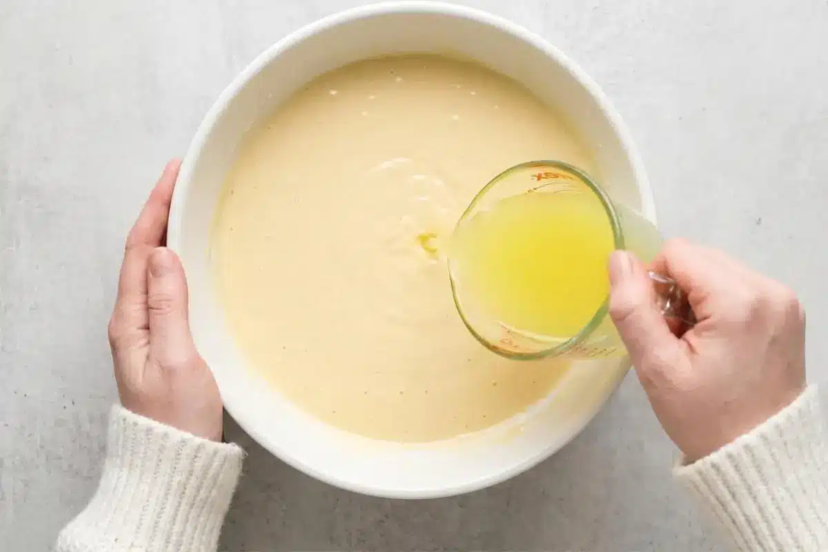 How to Make Limoncello Ricotta Cake A top-down view of a person wearing a cozy cream sweater pouring bright yellow limoncello from a glass measuring cup into a large white mixing bowl filled with creamy limoncello ricotta cake batter.