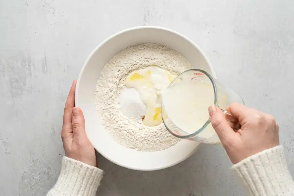 Mix the Wet into Dry For Raspberry Cake An overhead view of a baker wearing a cozy cream sweater pouring a light yellow liquid mixture from a glass measuring cup into a large white ceramic bowl containing the dry ingredients for a raspberry cake.