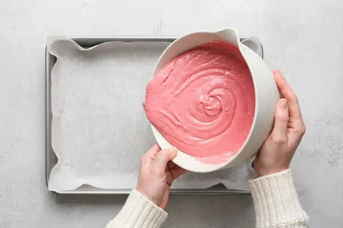 Pour and Bake For Raspberry Cake An overhead view of a baker wearing a cozy cream sweater pouring vibrant pink raspberry sheet cake batter from a large white ceramic bowl into a rectangular baking pan lined with parchment paper.