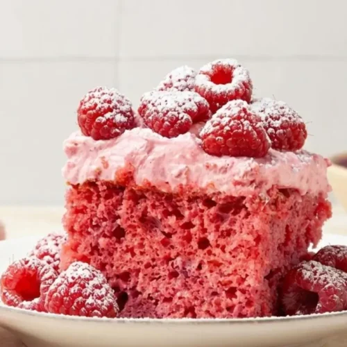 Nonna’s Simple Raspberry Cake (A Beautifully Messy, Foolproof Recipe) Raspberry Cake A close-up of a moist slice of pink raspberry cake topped with creamy pink frosting, fresh raspberries, and a light dusting of powdered sugar, served on a rustic white plate.