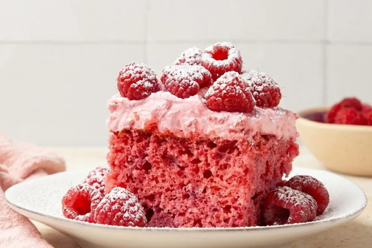 Nonna’s Simple Raspberry Cake (A Beautifully Messy, Foolproof Recipe) Raspberry Cake A close-up of a moist slice of pink raspberry cake topped with creamy pink frosting, fresh raspberries, and a light dusting of powdered sugar, served on a rustic white plate.