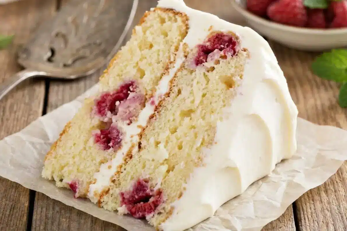 Nonna’s Foolproof Raspberry Lemon Cake: A Sunny, No-Sink Italian Bake Raspberry Lemon Cake A generous, comforting slice of homemade raspberry lemon cake with fluffy white frosting, served casually on parchment paper over a weathered wooden table next to a vintage silver cake server.