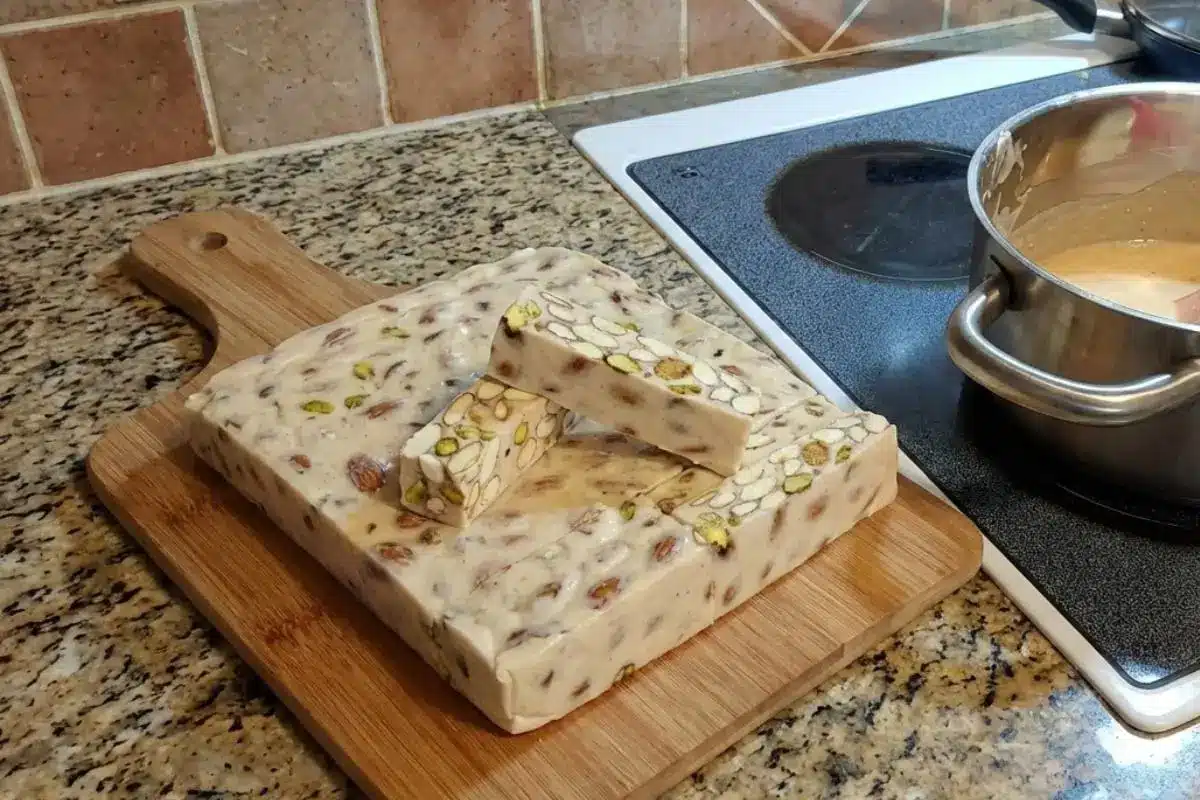 Rena's No-Fail Candy Tips For Your Classic Italian Torrone A freshly made block of classic Italian torrone packed with pistachios and almonds, resting on a wooden cutting board next to a pot of warm syrup on a home stovetop.