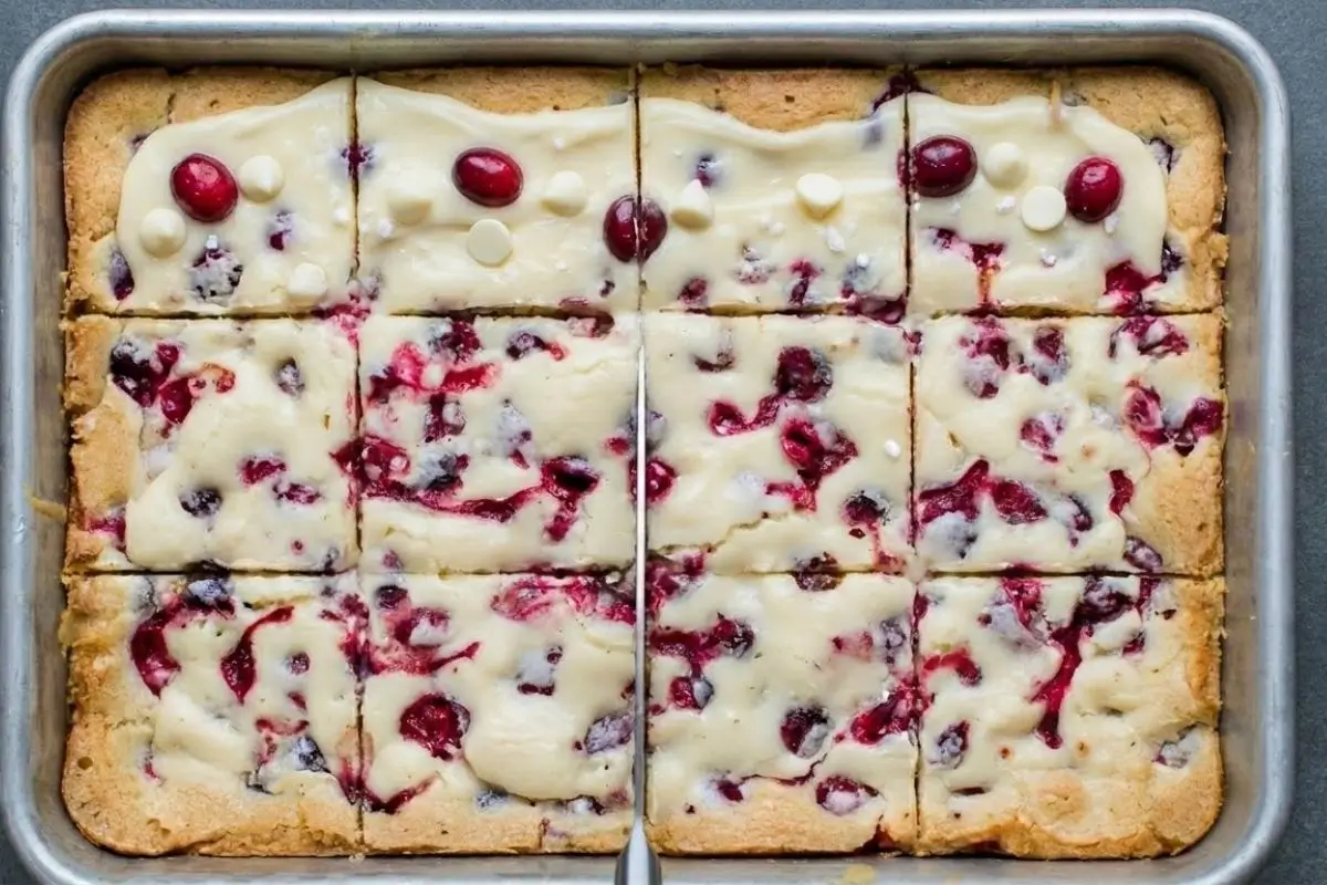 Rena's No-Fail Tips For Cranberry Cookie Bars A silver baking pan of cranberry cookie bars being sliced into even squares with a metal knife, showing the vibrant red cranberry swirls and white chocolate topping.