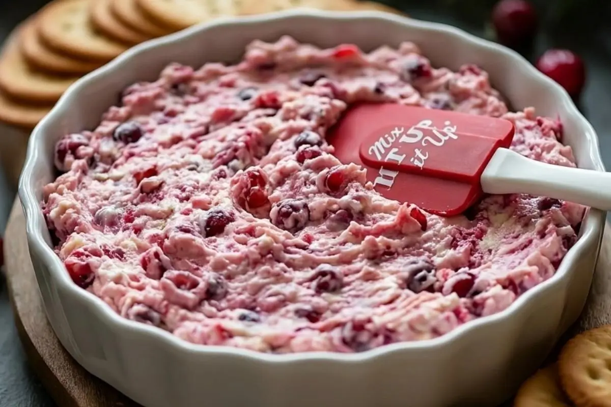Rena's No-Fail Tips For Your Baked Cranberry Cream Cheese Dip A bright red spatula spreading the unbaked creamy, pink mixture of a baked cranberry cream cheese dip into a rustic white fluted baking dish, surrounded by buttery crackers.