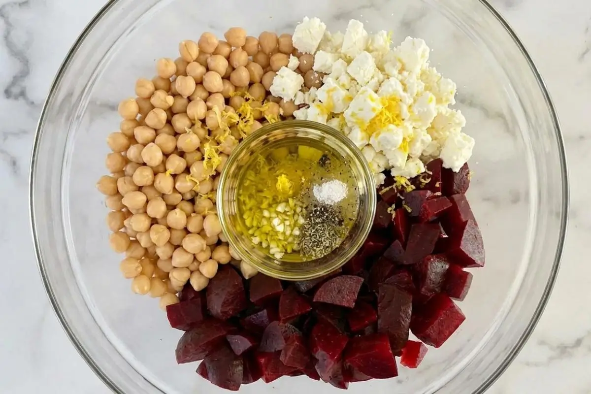Rena's No-Fail Tips For Your Chickpea Salad with Feta A top-down view of a clear glass bowl containing neatly arranged sections of chickpeas, diced beets, and crumbled cheese, with a small jar of olive oil, garlic, and herb dressing resting in the center to illustrate no-fail tips for a beet and chickpea salad with feta.